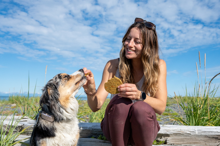 a woman stiting on a log in a field feeding freeze-dried food as a treat to a dog