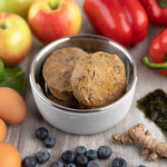 A top-down view of a stainless steel dog bowl filled with Artisan Raw freeze-dried food. Fresh, whole ingredients like raw eggs and vibrant vegetables are scattered around the dish to showcase the natural color and freshness of the recipe.