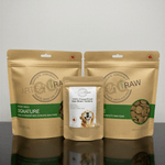 Two Artisan Raw Signature freeze-dried dog food bags and a bag of Bison Tendons treats displayed together on a reflective black surface.