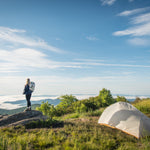 A person stands on a rock near a tent, overlooking a mountainous landscape with clouds below and a blue sky above.