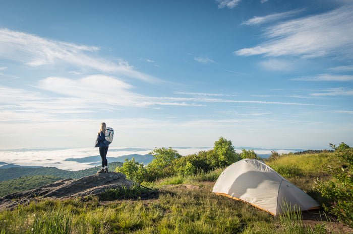 A person stands on a rock near a tent, overlooking a mountainous landscape with clouds below and a blue sky above.